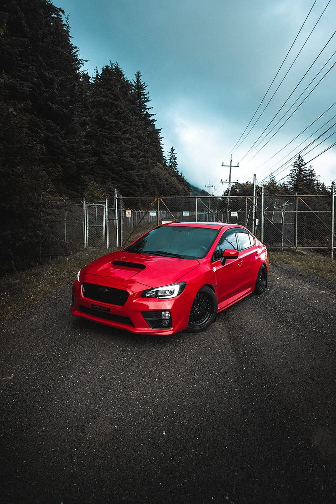 Red Subaru WRX STI with black wheels parked on a gravel road beside a metal fence and tall green trees.