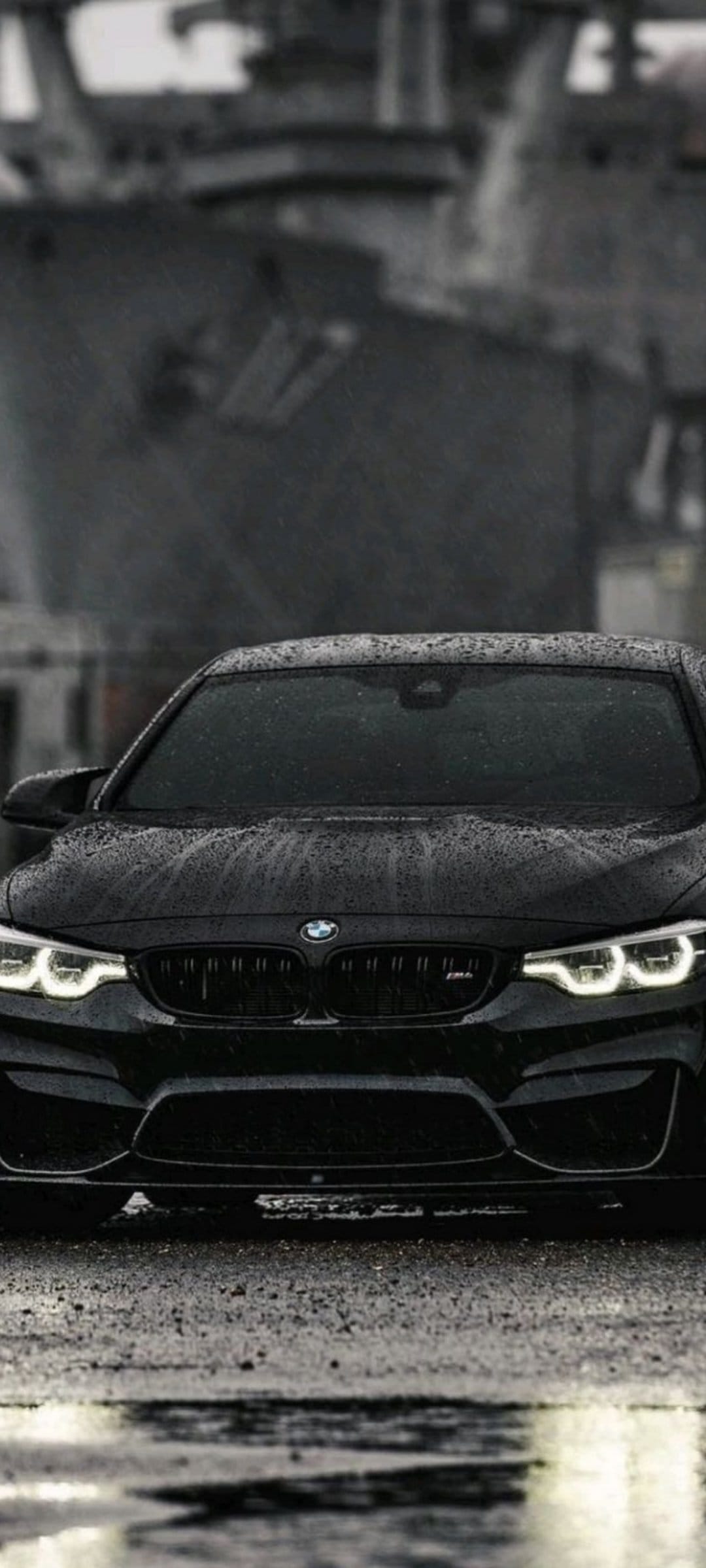 Black BMW M4 coupe parked on a wet asphalt road at night with glowing headlights and rain droplets on the hood.