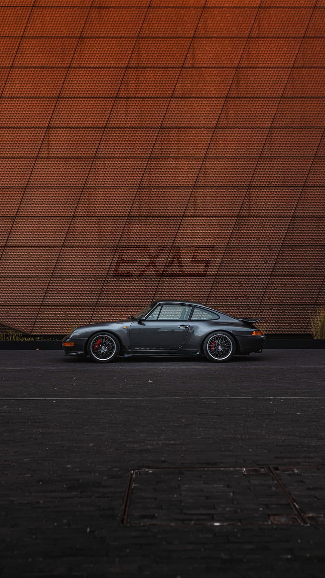 Classic grey Porsche 911 parked on dark cobblestones in front of a red brick wall with an etched EXAS logo.
