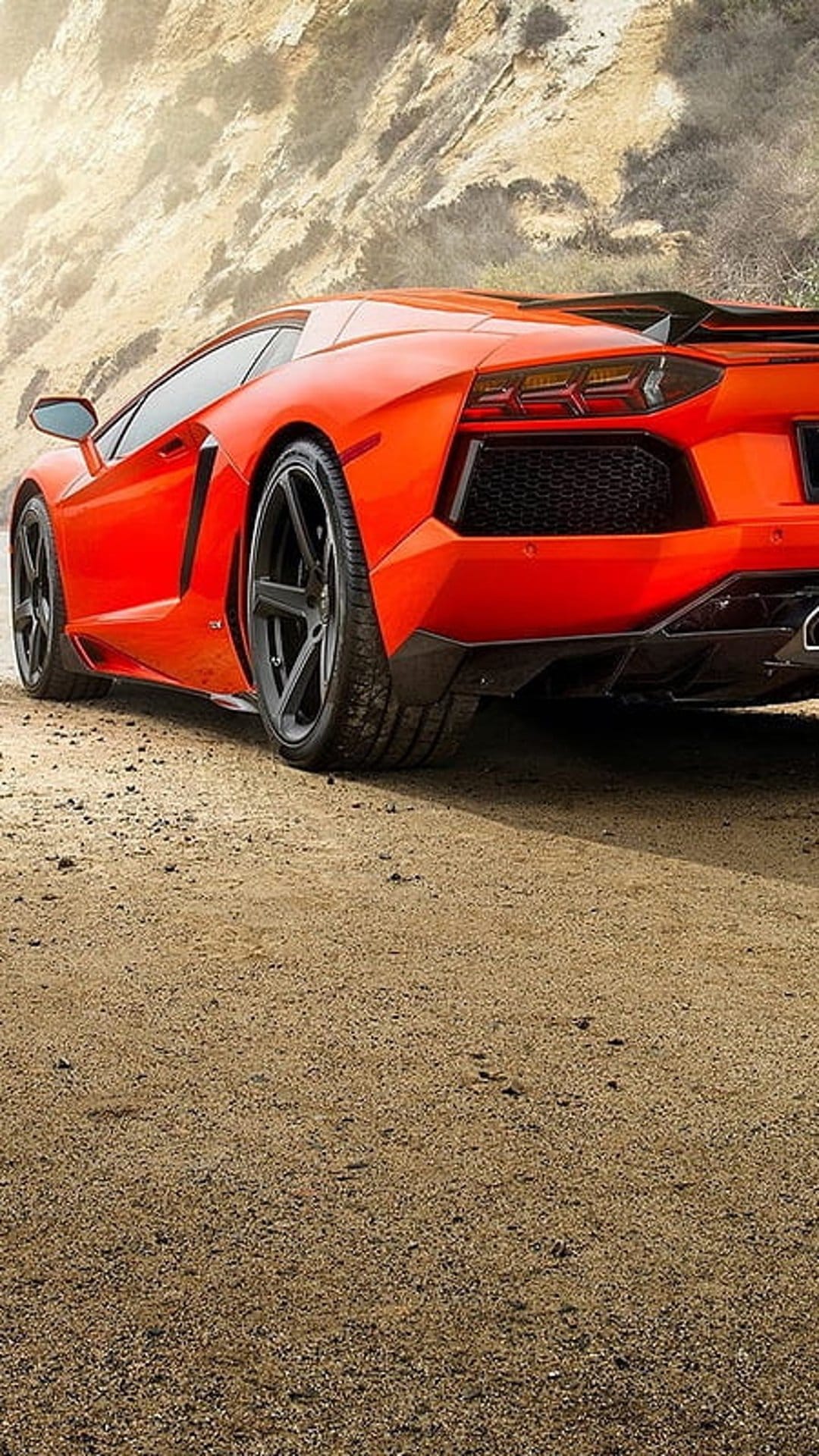 Orange Lamborghini Aventador rear view on a desert road with rugged sandstone rocks and glowing red taillights.