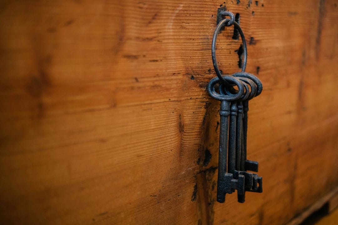 Antique skeleton keys hang from a heavy iron ring against a weathered wooden chest showing rusted metal textures.