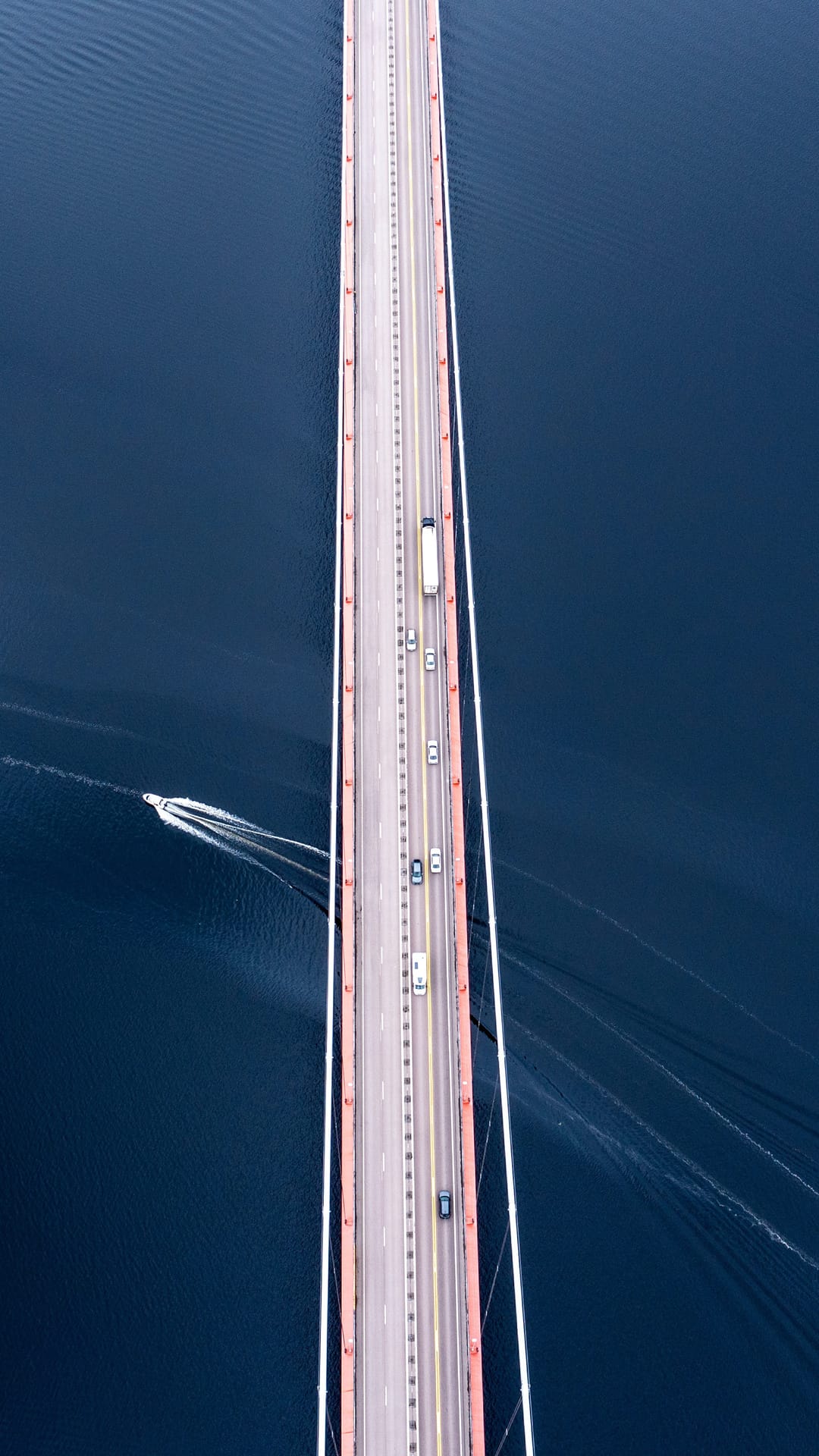 Modern bridge with red supports crosses deep blue water with cars and a white speedboat wake seen from above.