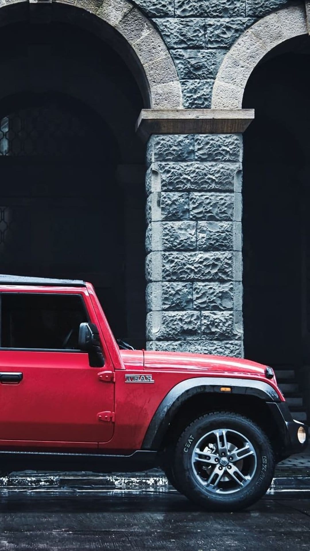 Red Mahindra Thar SUV parked next to a textured stone archway under soft outdoor lighting conditions.