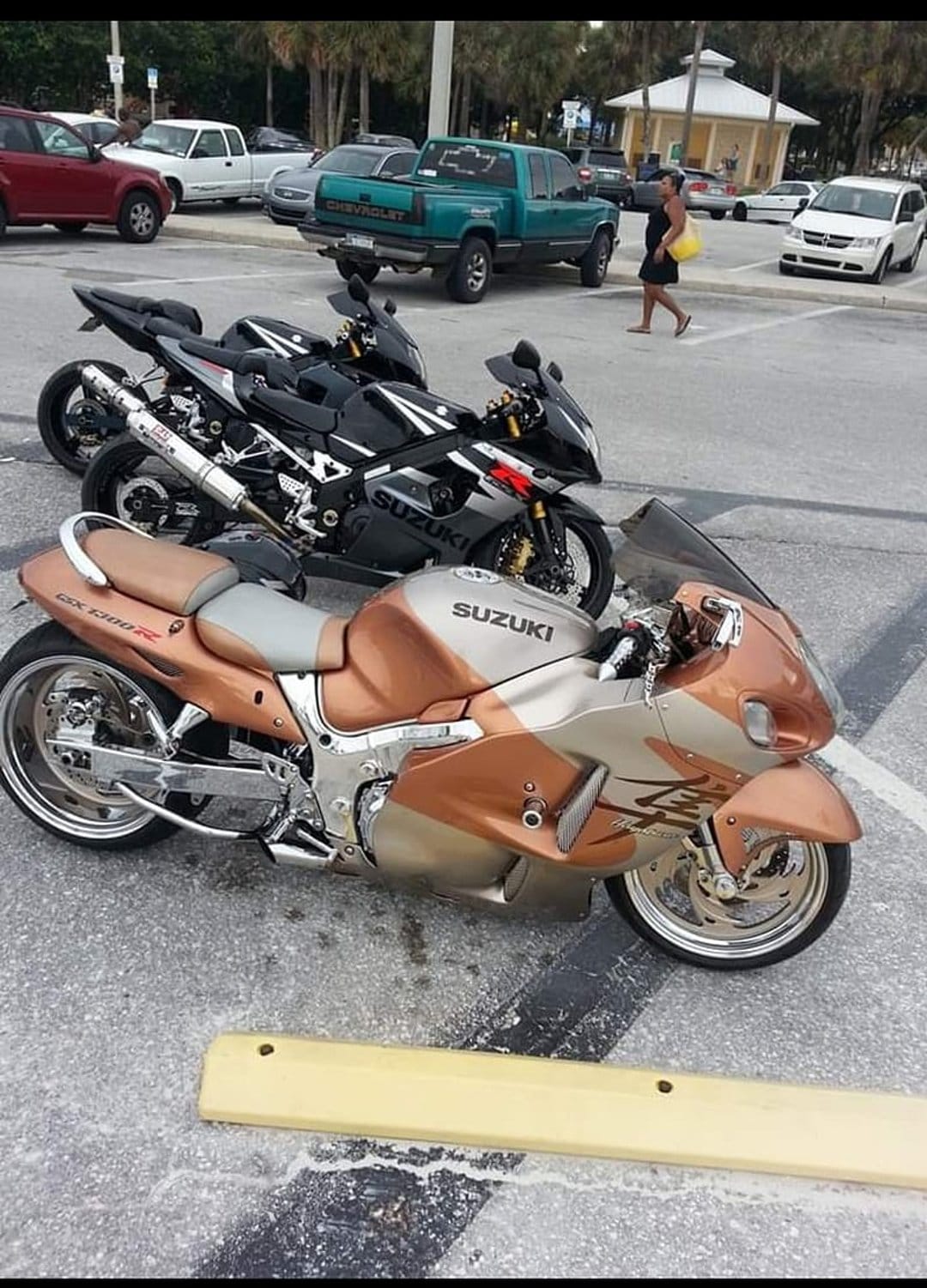 Suzuki Hayabusa with gold fairings parked next to a black GSXR under palm trees in a sunny outdoor lot.