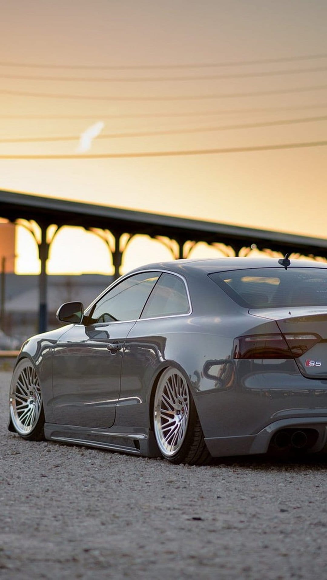 Audi S5 coupe with custom wheels and lowered suspension parked at sunset against an industrial backdrop.