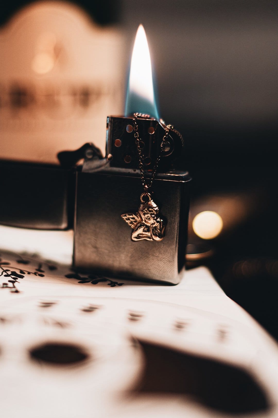 Chrome Zippo lighter with a flickering orange flame and a small gold angel charm hanging from a metal chain.