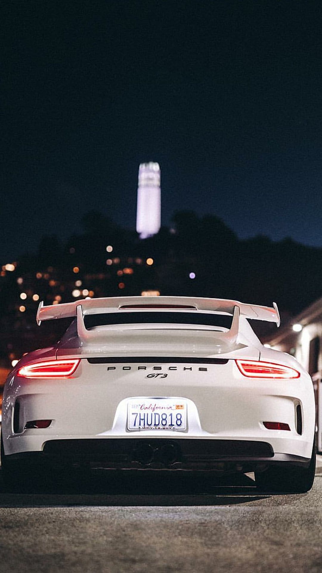 White Porsche GT3 rear view with glowing red taillights parked on an urban street with city tower lights at night.