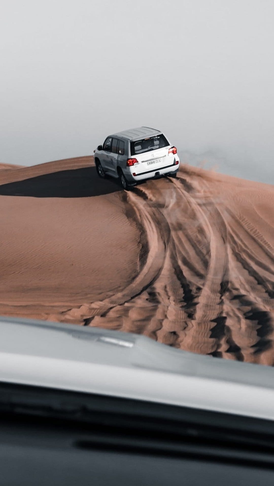 White SUV climbing a steep sandy ridge leaving deep tire tracks under bright desert sunlight in a vertical view.