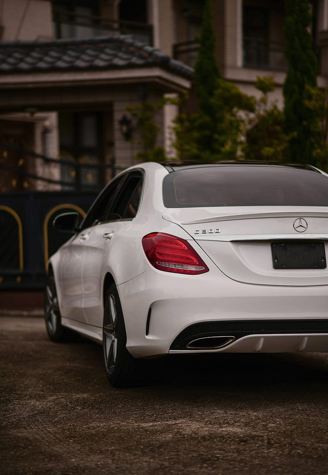 White Mercedes-Benz C-Class sedan with a black sunroof parked on a gravel drive before an ornate luxury mansion.