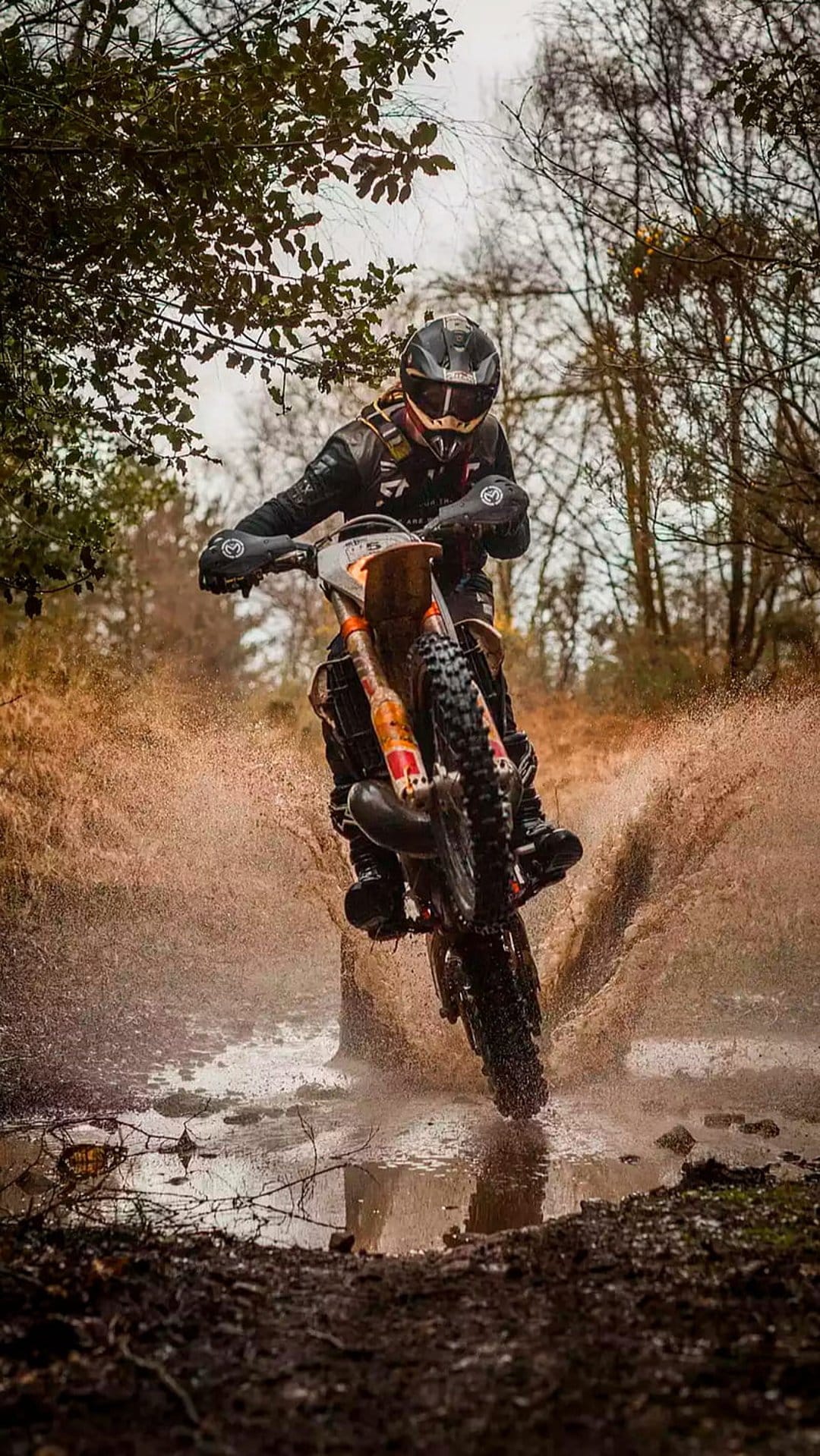 Motocross rider in gear jumps through a deep puddle creating a dramatic spray of mud and water in a forest.