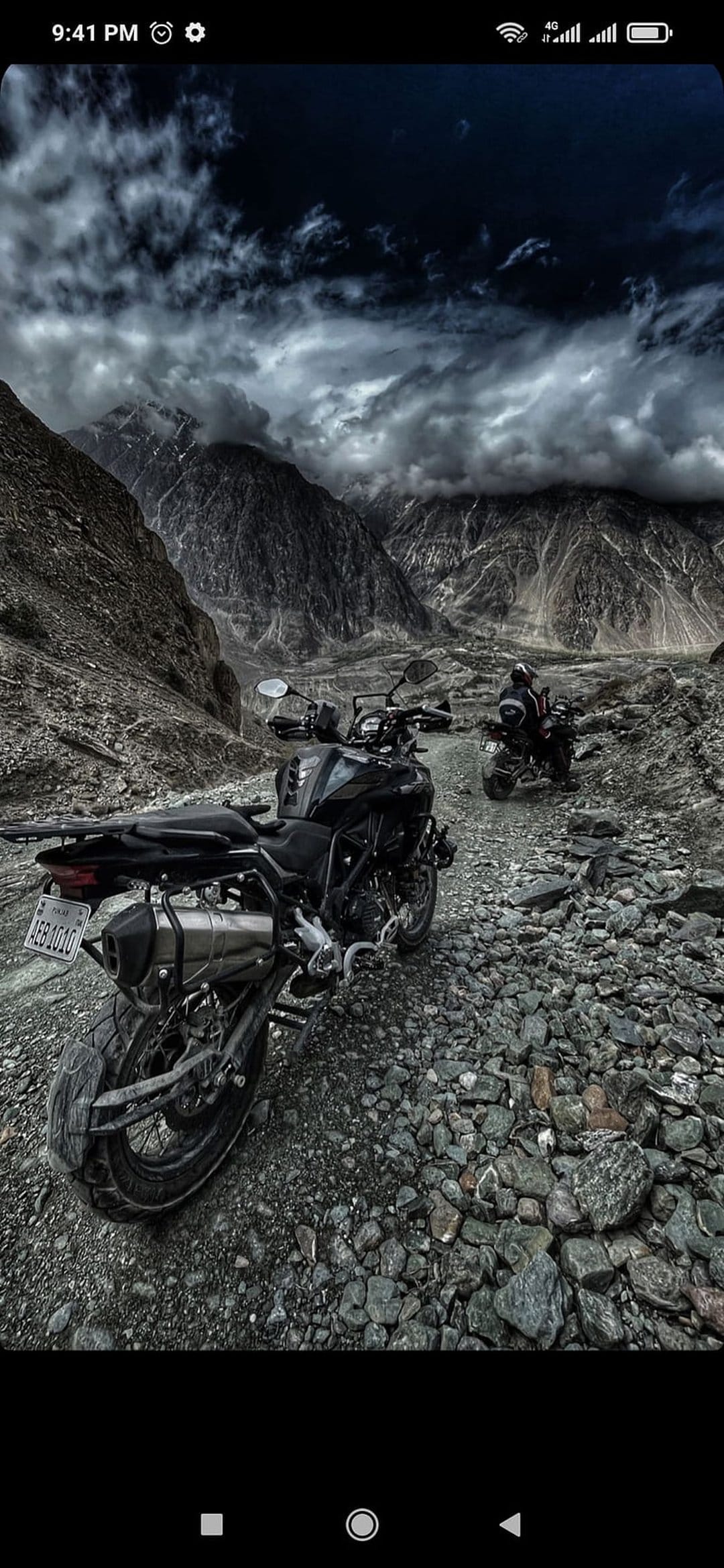 Two adventure motorcycles on a rocky mountain ridge under a dark stormy sky with jagged peaks in the background.