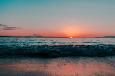 Foamy ocean waves wash over a dark sandy beach under a glowing orange sky with silhouetted land in the distance.