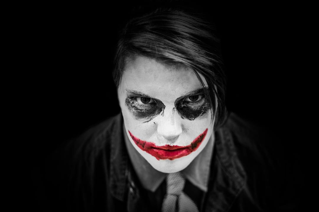 Joker-style face paint with a red smile and white skin against a black background in a moody close-up portrait.