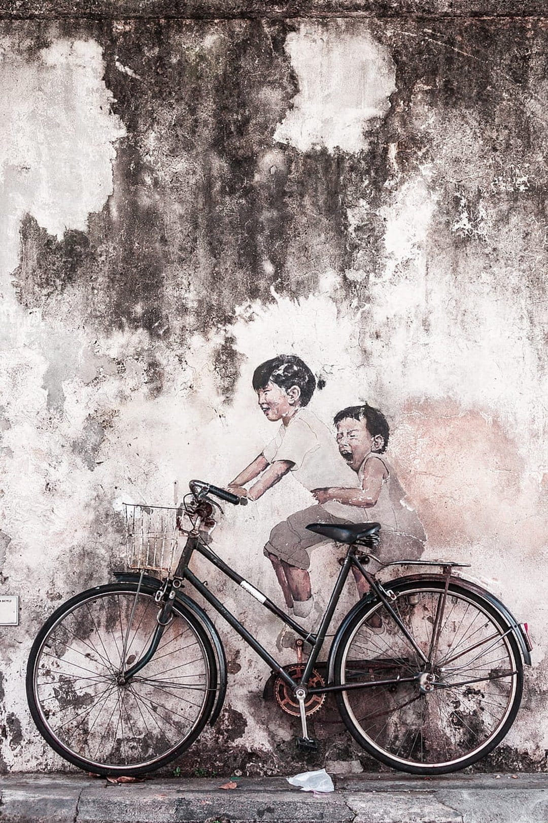 Two children ride a bicycle in a street art mural on a cracked, weathered wall with visible texture and peeling paint.