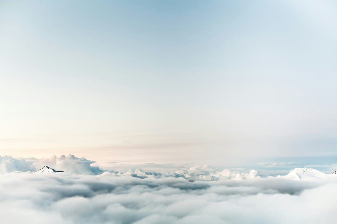 Snow-covered mountain summits piercing through thick white clouds under a soft blue and orange gradient sky.