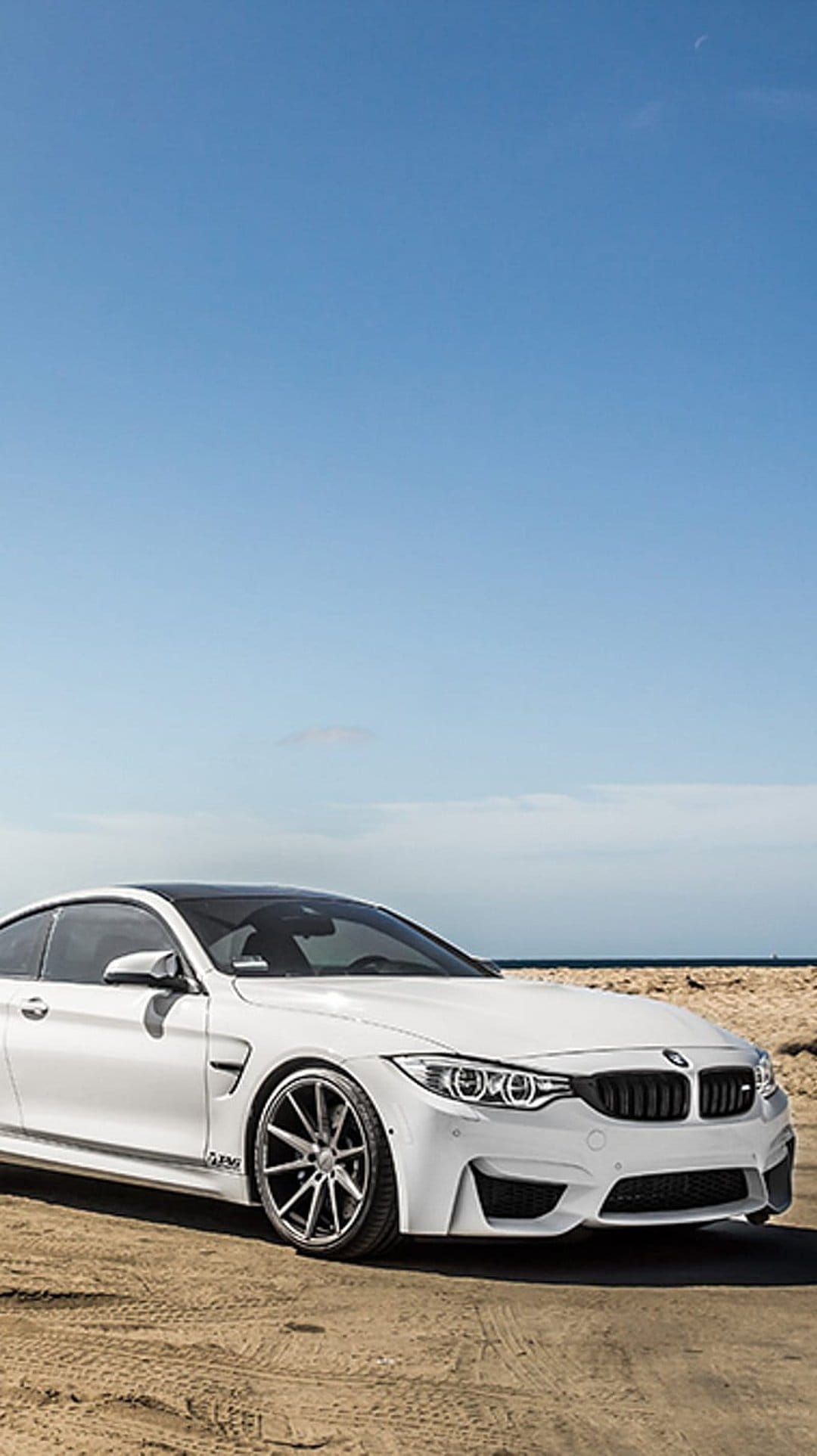 White BMW M4 coupe parked on golden beach sand with a clear blue sky and calm ocean waves in the background.