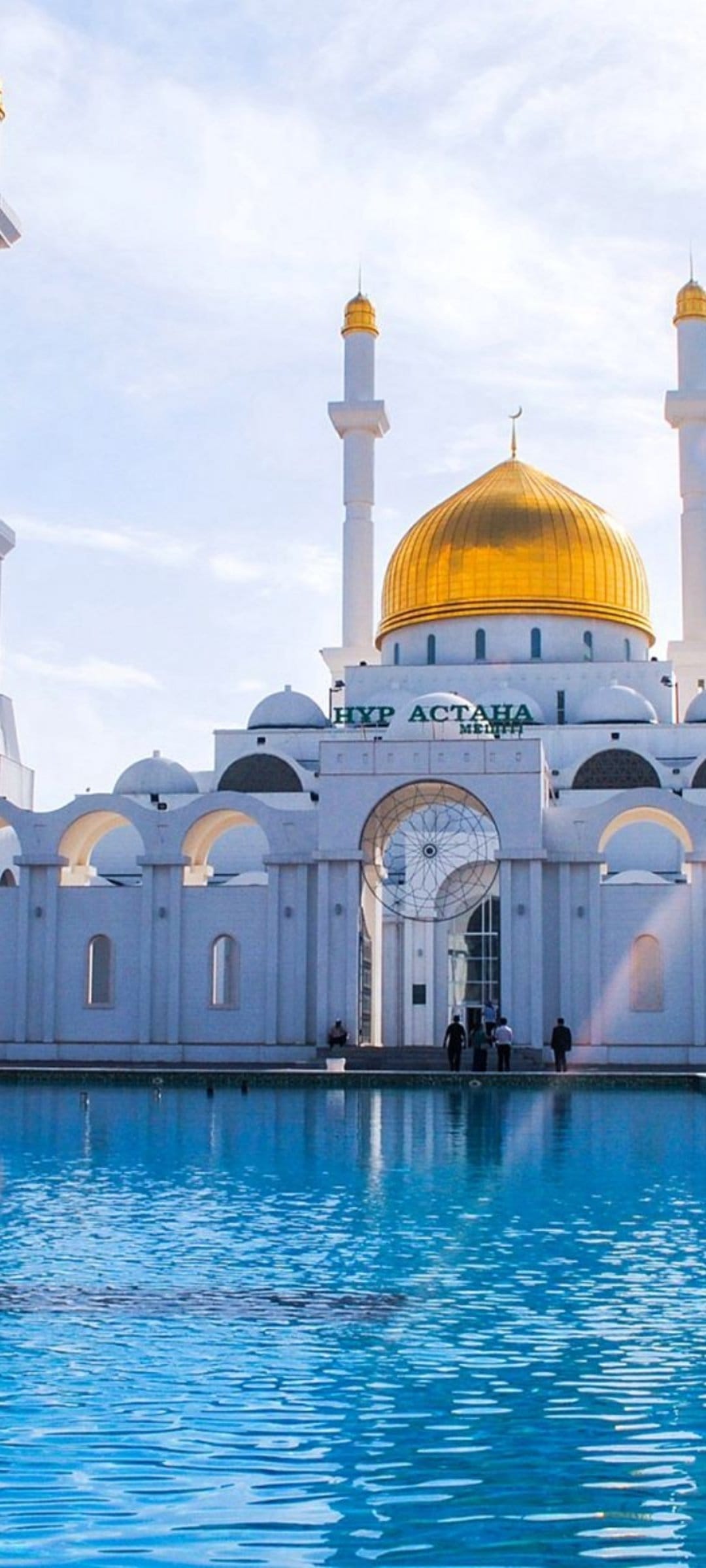 Nur Astana Mosque with a gold dome and white minarets reflects in a blue pool under a bright sunny sky.