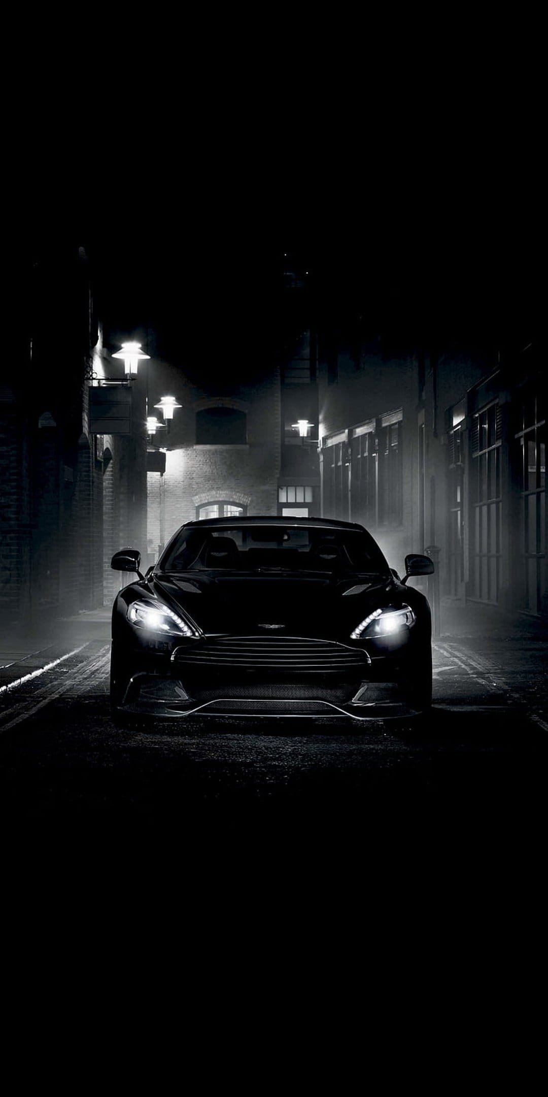 Black Aston Martin sports car parked in a dark rainy alleyway between brick buildings under amber streetlights.