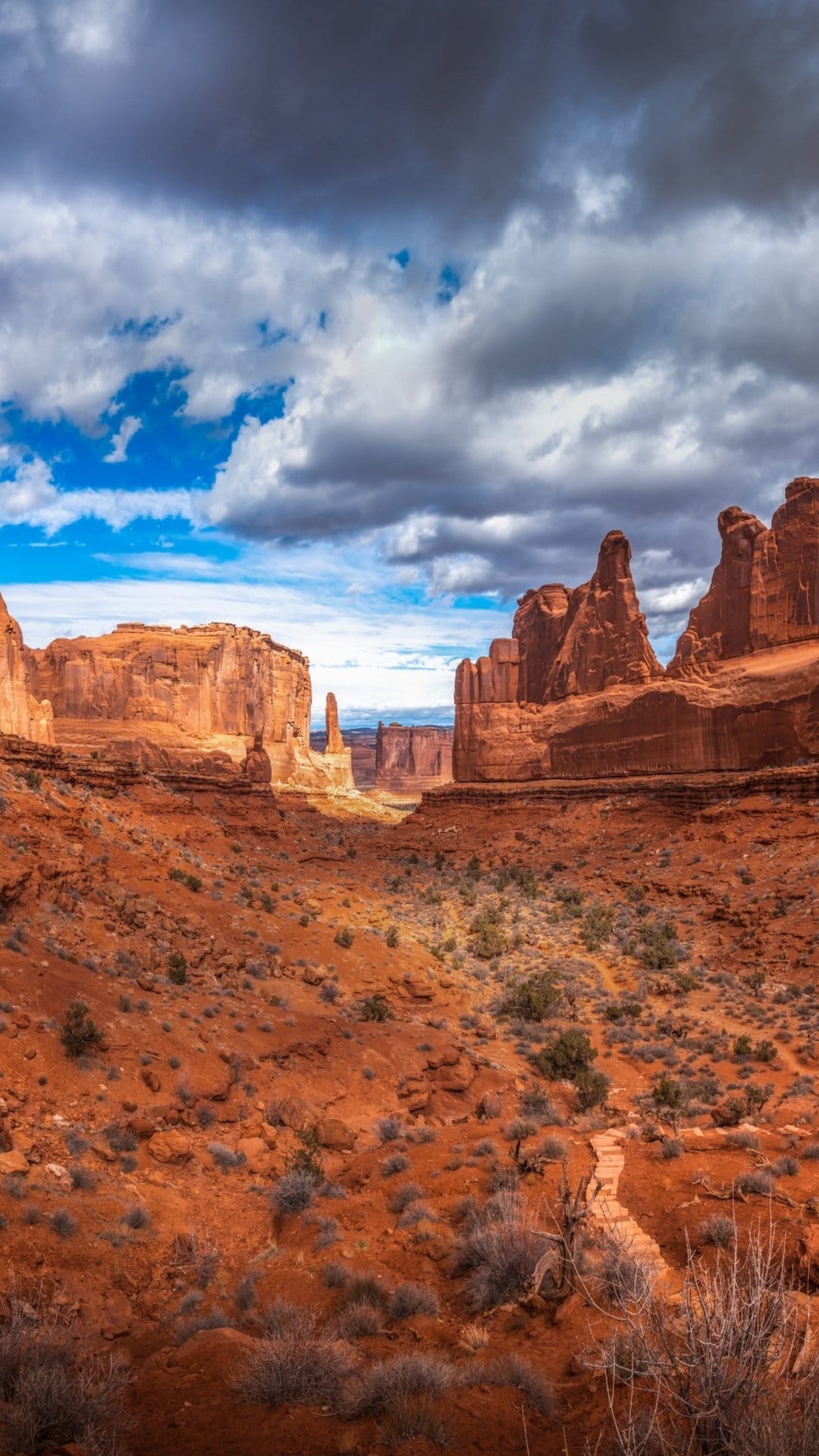 Red rock canyons and a desert path sit beneath a blue and stormy sky during a vibrant golden hour sunset.