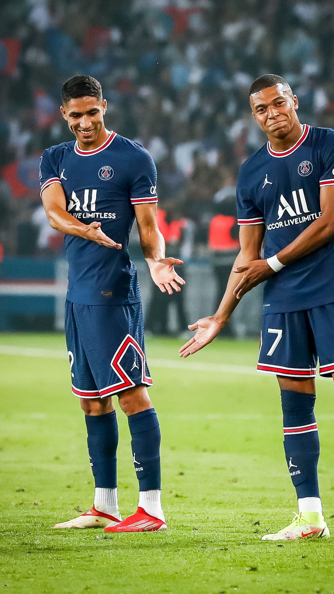 Kylian Mbappé and Achraf Hakimi celebrate on a green football pitch wearing navy blue Paris Saint-Germain kits.