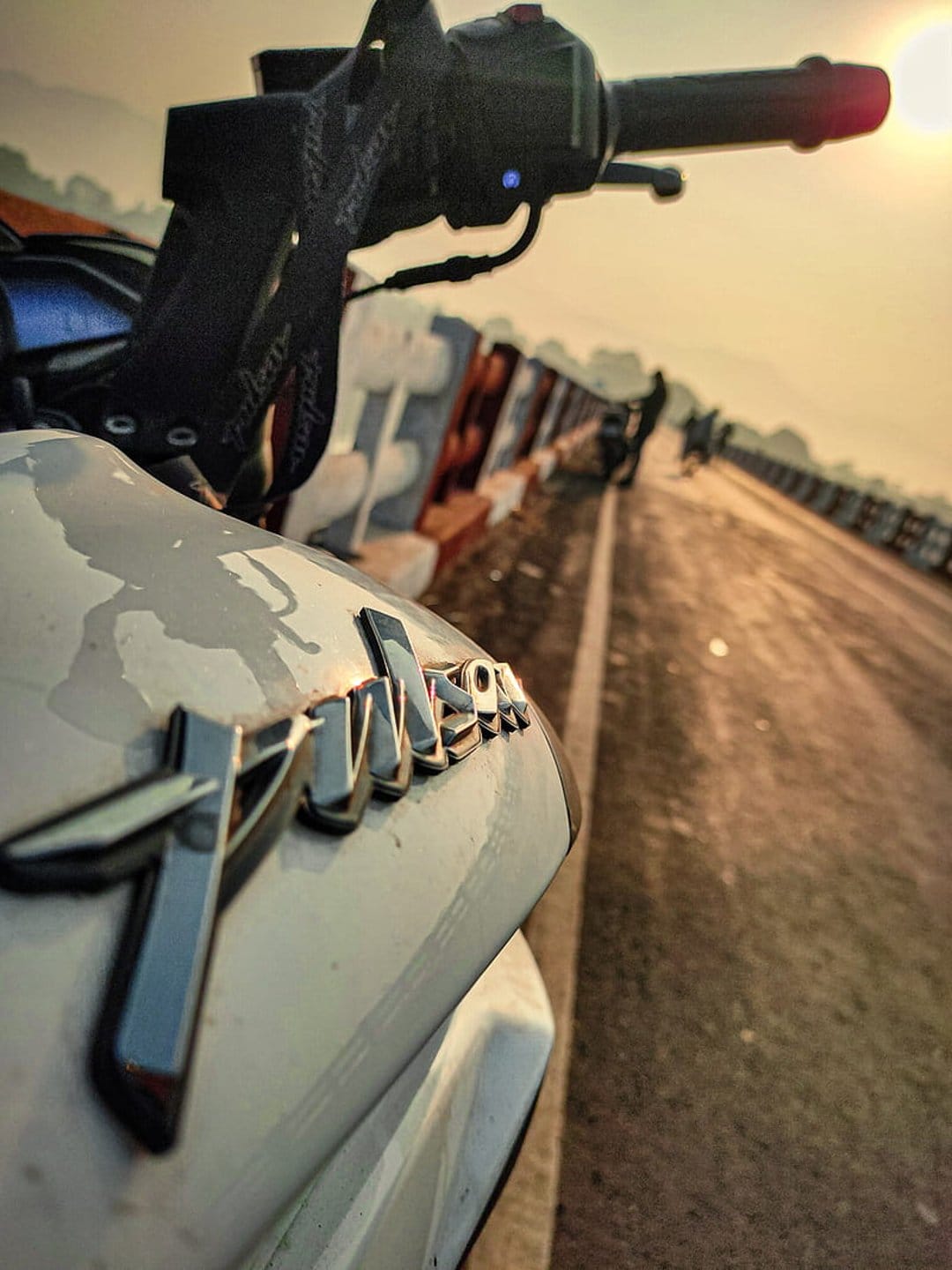 White motorcycle emblem and handlebars on a sunset bridge, featuring warm lighting and a blurred roadway.
