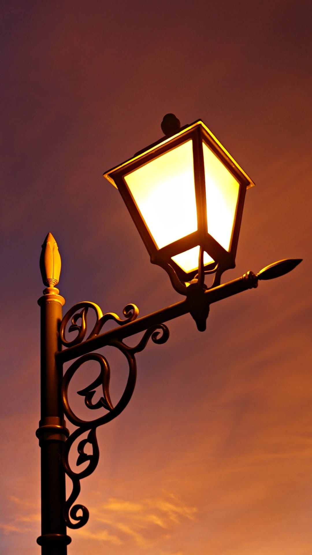 Ornate wrought iron street lamp glows with warm light against a gradient sky of purple and orange clouds at dusk.