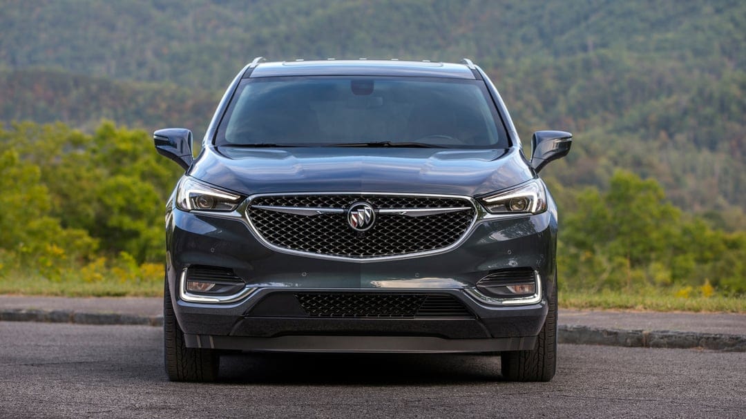 Front view of a dark grey Buick SUV parked on an asphalt road with a blurred forest mountain background.