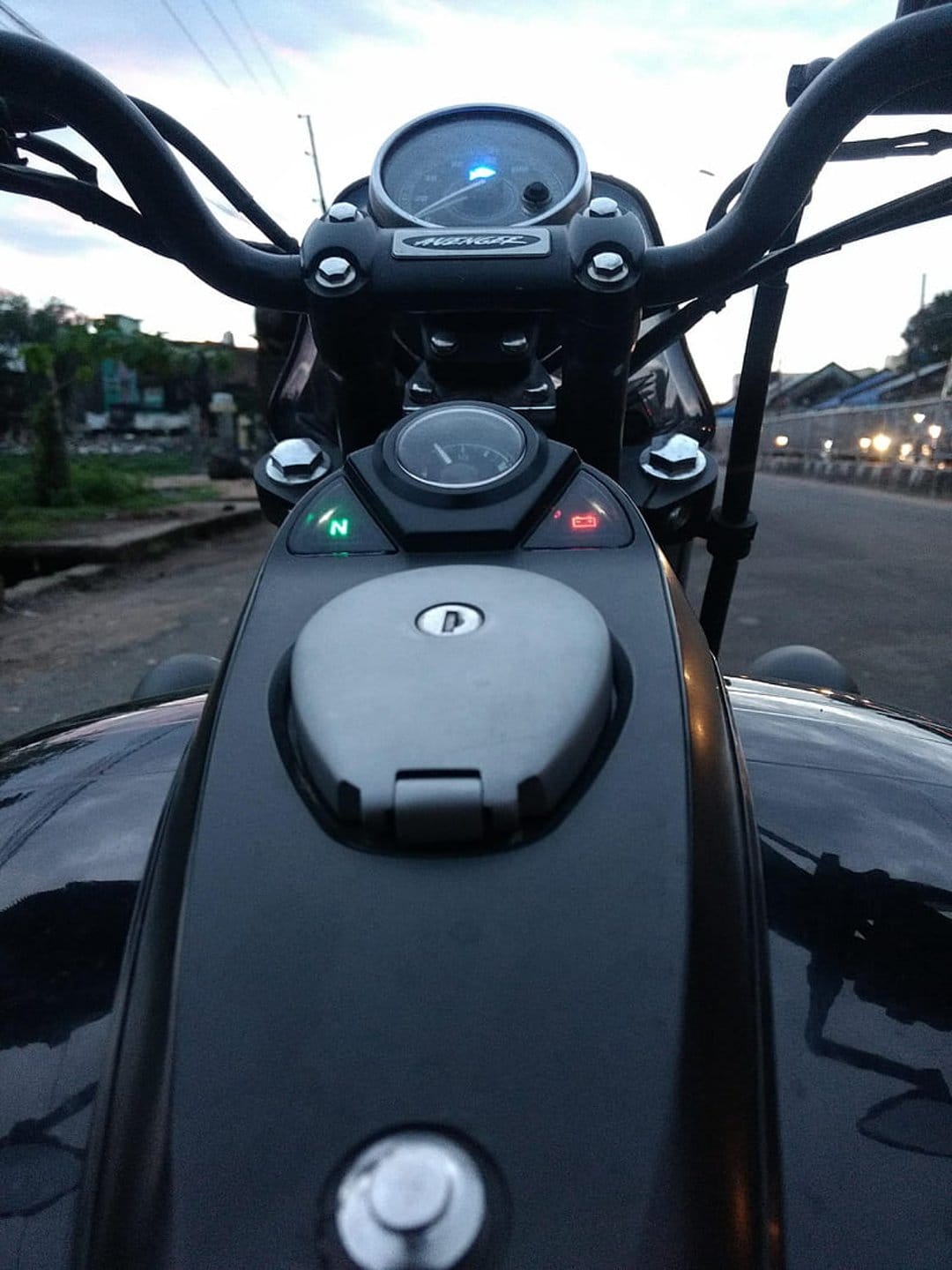 Motorcycle speedometer glows blue next to chrome handlebars and a glossy fuel tank during the evening twilight.