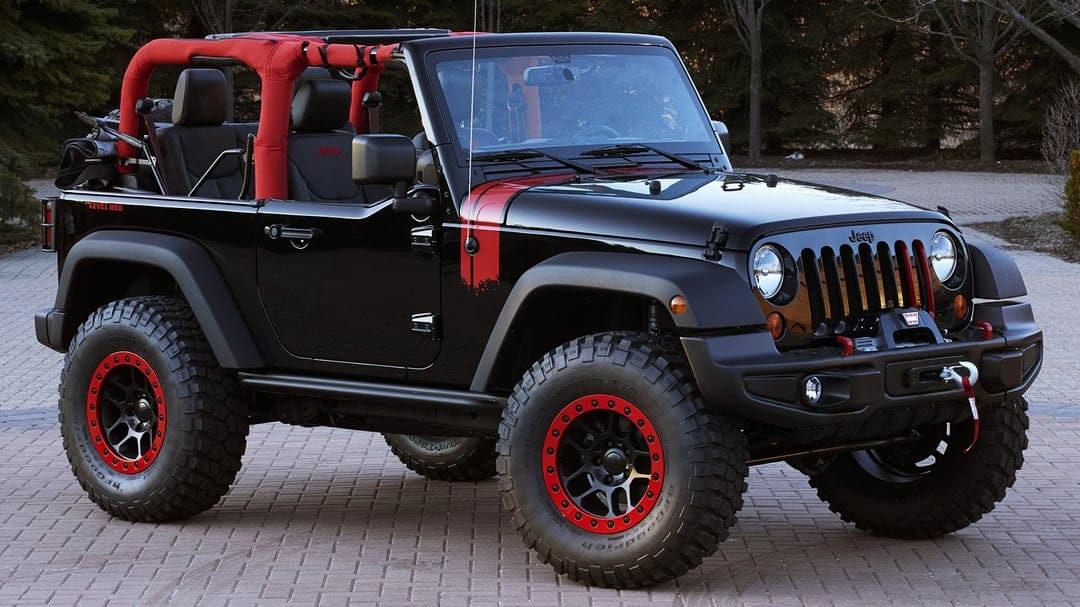 Side profile of a black Jeep Wrangler with red roll bars and oversized off-road tires parked on a brick driveway.