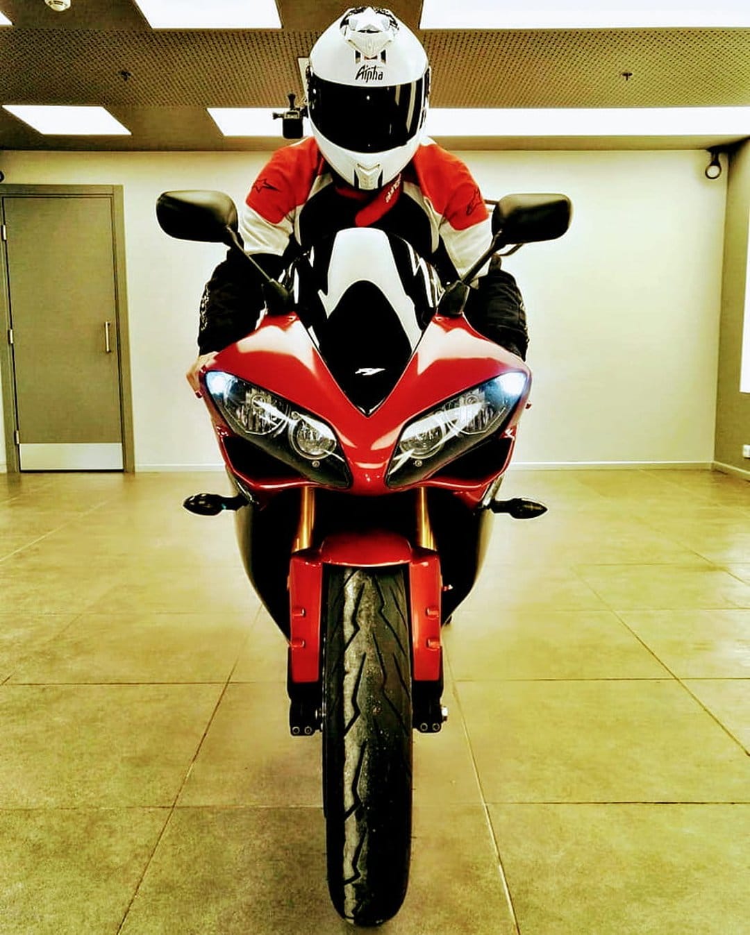 Motorcyclist in a white and red helmet sits on a red sportbike over a tiled floor in a vertical composition.