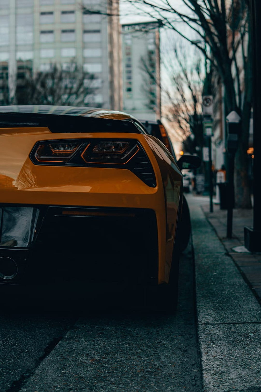 Yellow sports car rear tail lights glow against a blurred city street with soft bokeh lights and urban buildings.