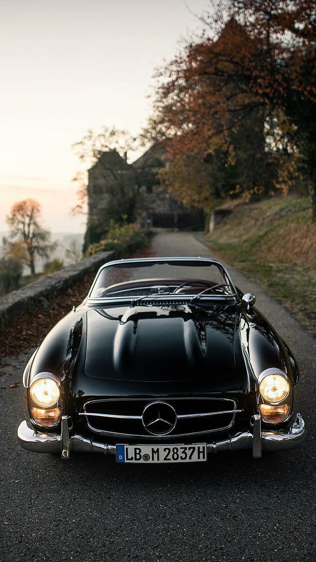 Black vintage Mercedes convertible driving on a tree-lined road with autumn leaves and a stone building behind.