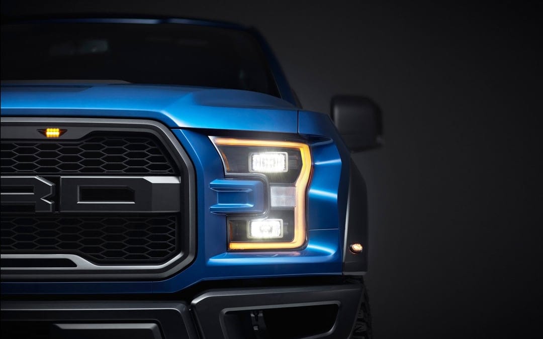 Front corner of a blue Ford Raptor with glowing C-shaped amber headlights and white lamps in a dark studio.
