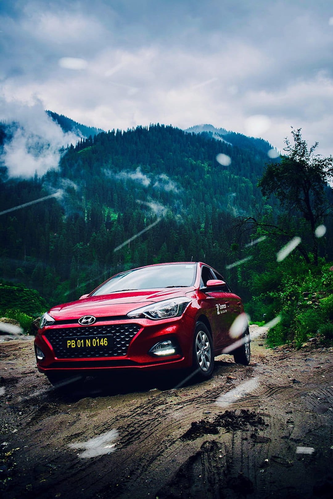 Red Hyundai i20 drives through a muddy mountain road surrounded by mist-covered pine trees and cloudy skies.