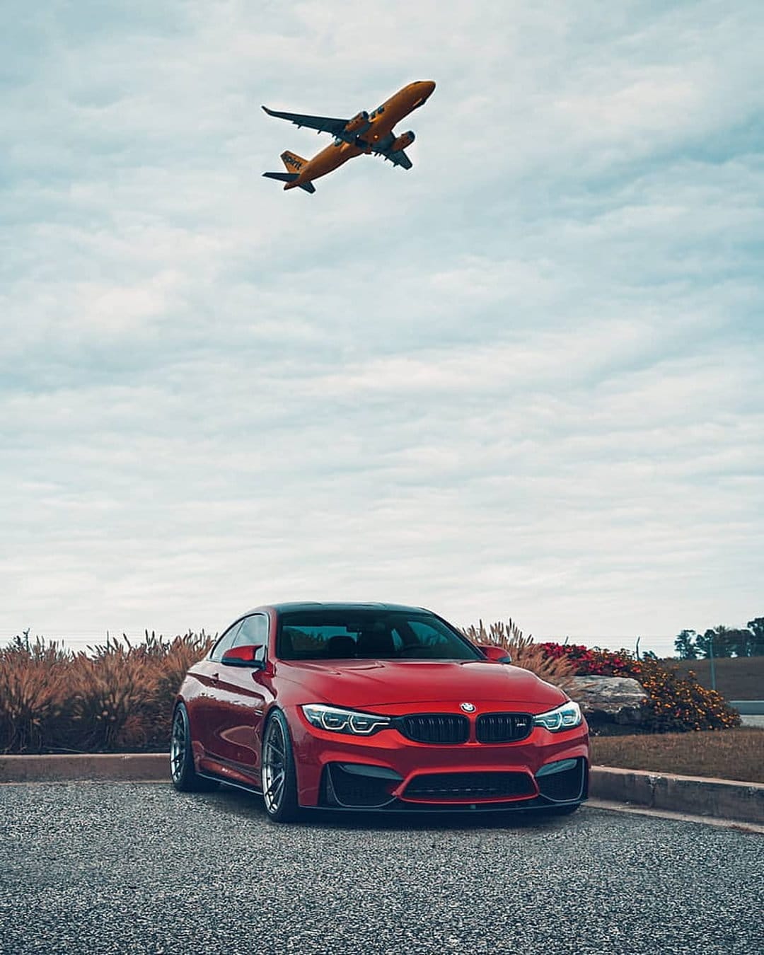 Red BMW M4 coupe parked on a gravel lot while a vibrant orange airplane flies overhead under a cloudy sky.