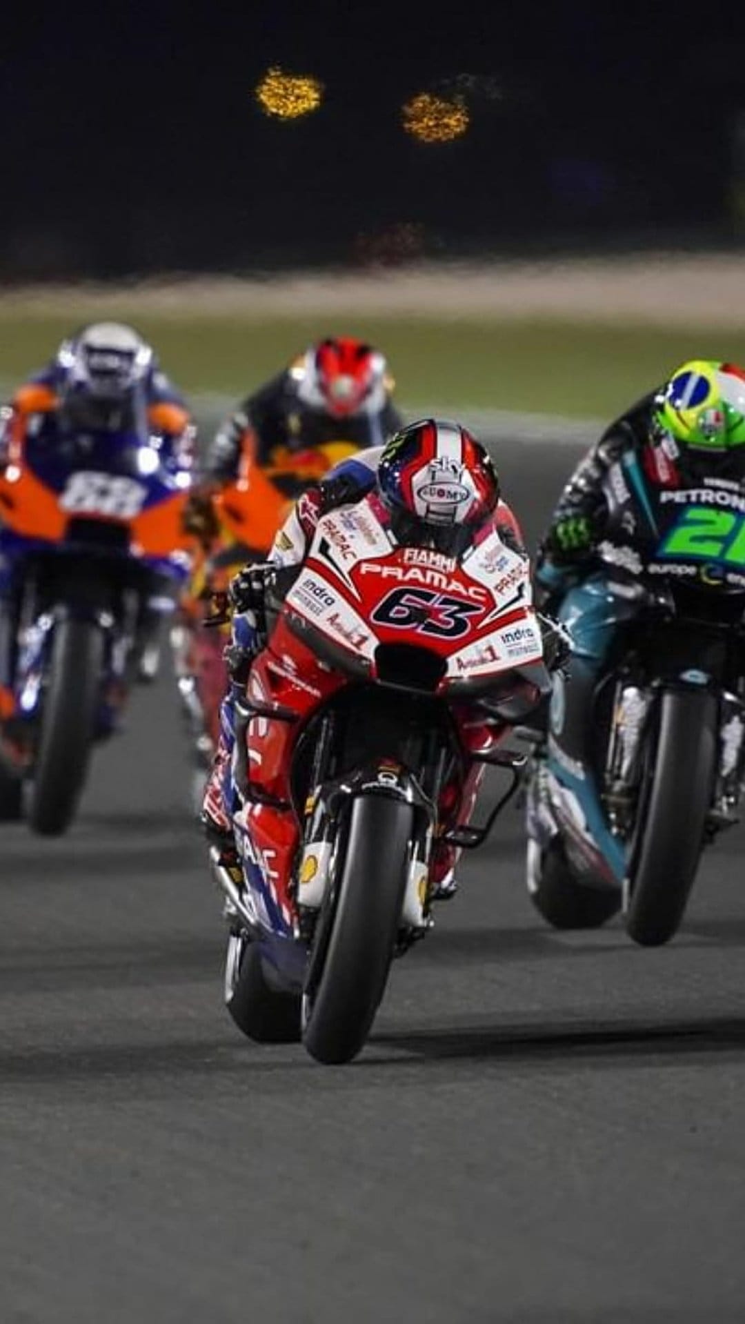 Professional motorcycle rider wearing number 63 leans into a turn on a lit circuit at night during a race.
