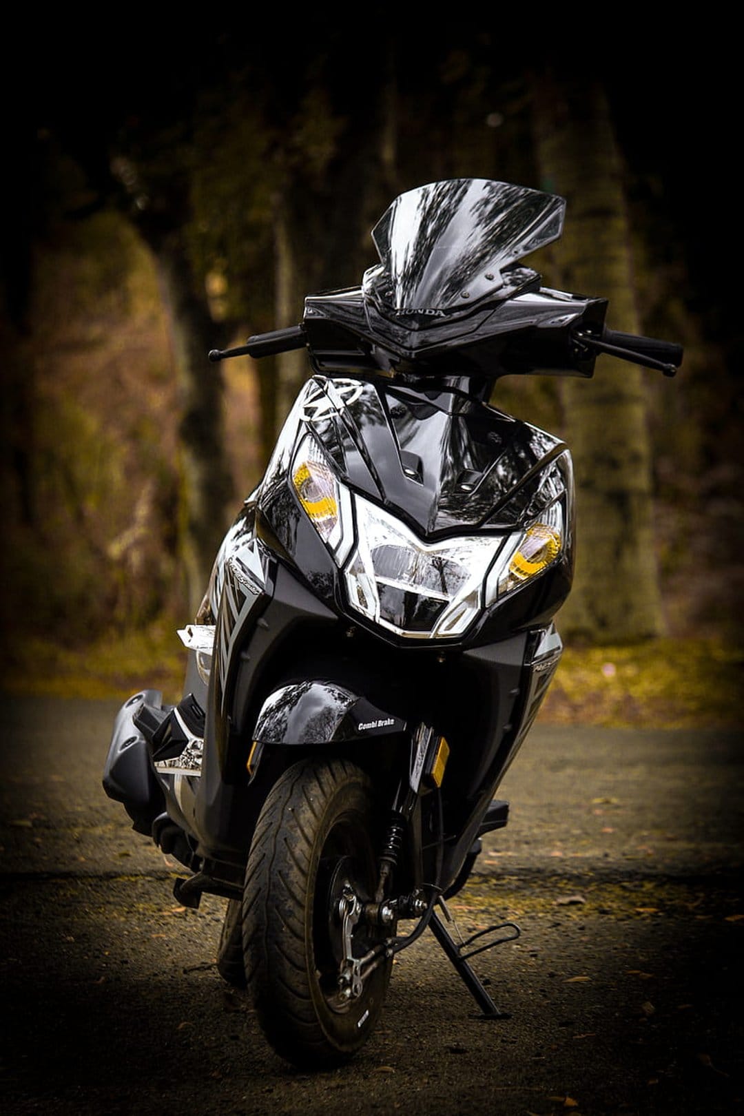 Black scooter with yellow headlight accents parked on a dirt path in a blurred forest, low-angle view.
