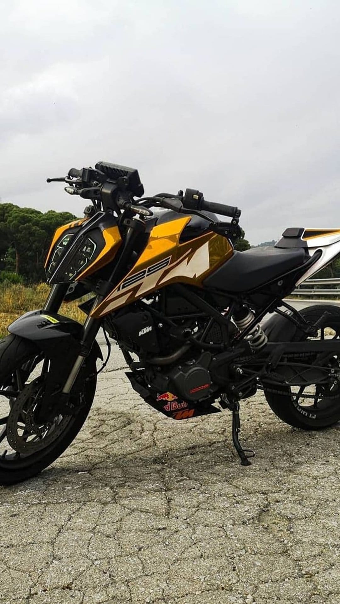 KTM Duke 125 motorcycle with gold graphics parked on a cracked road under a cloudy sky with blurred trees.