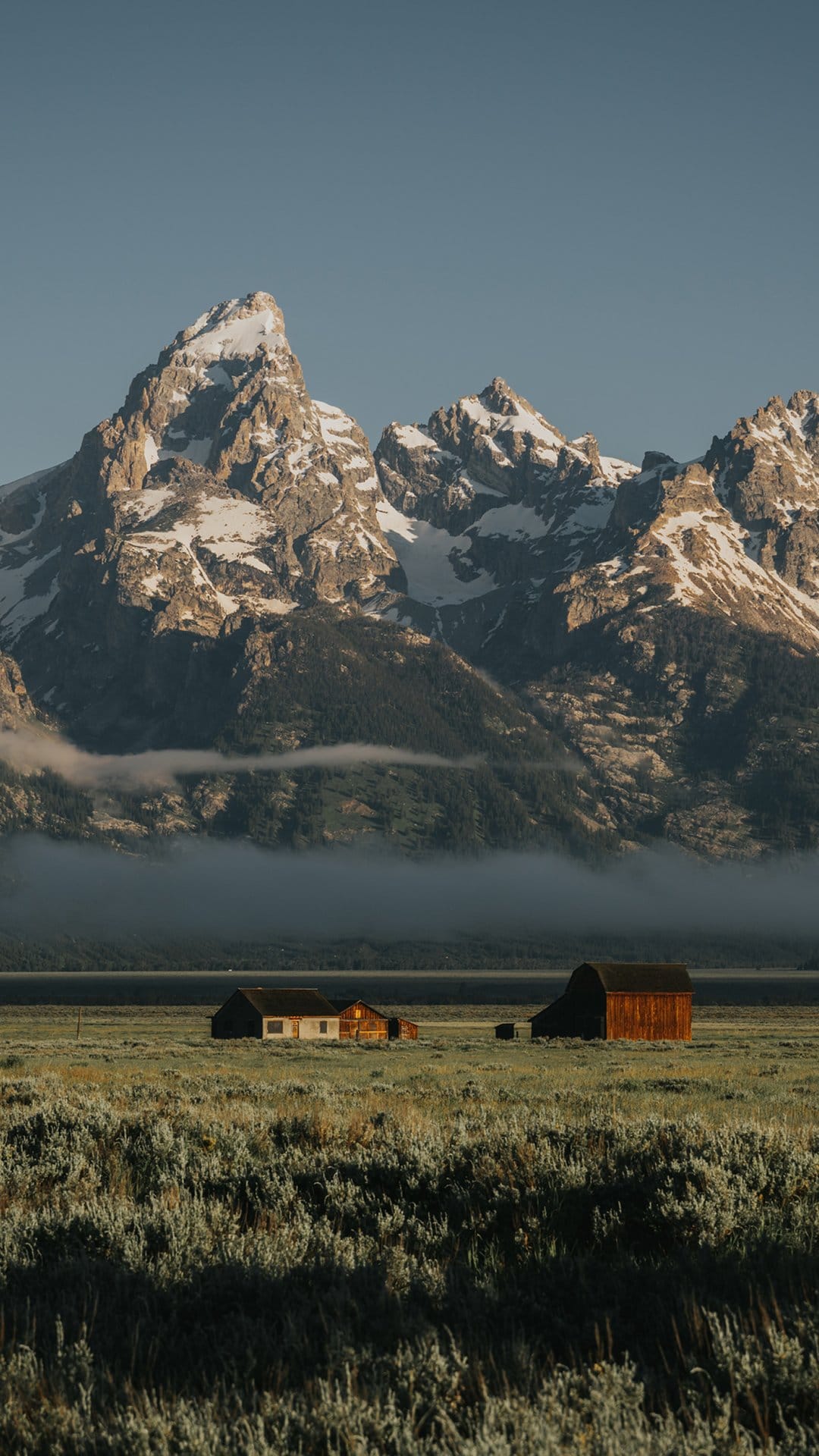 Weathered wooden barns stand in a green field before the jagged, snow-dusted peaks of the Grand Teton mountains.