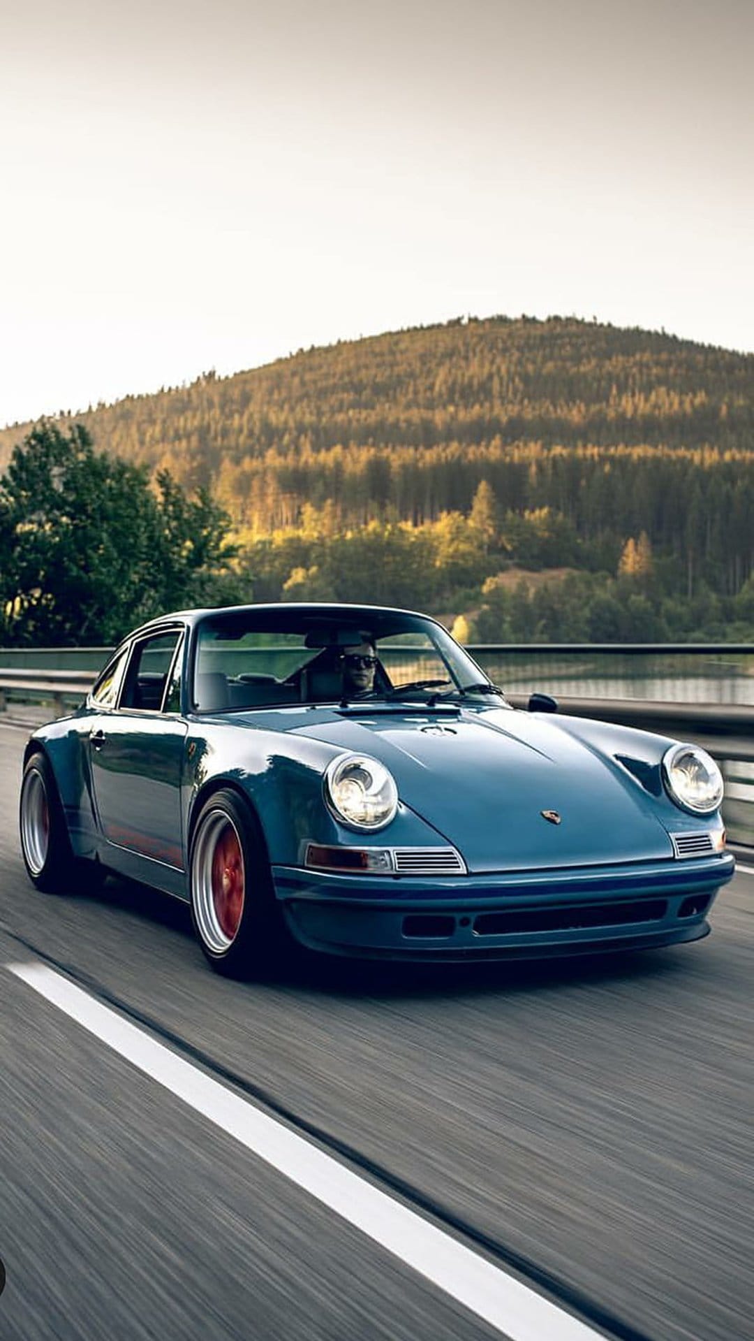 Blue vintage Porsche 911 drives on a paved forest road with sunlight streaming through tall evergreen trees.