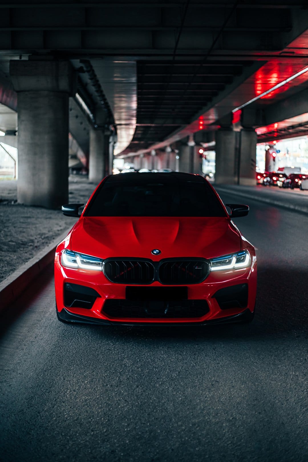 Red BMW M5 sport sedan with bright headlights parked beneath a concrete bridge in a dark urban city setting.