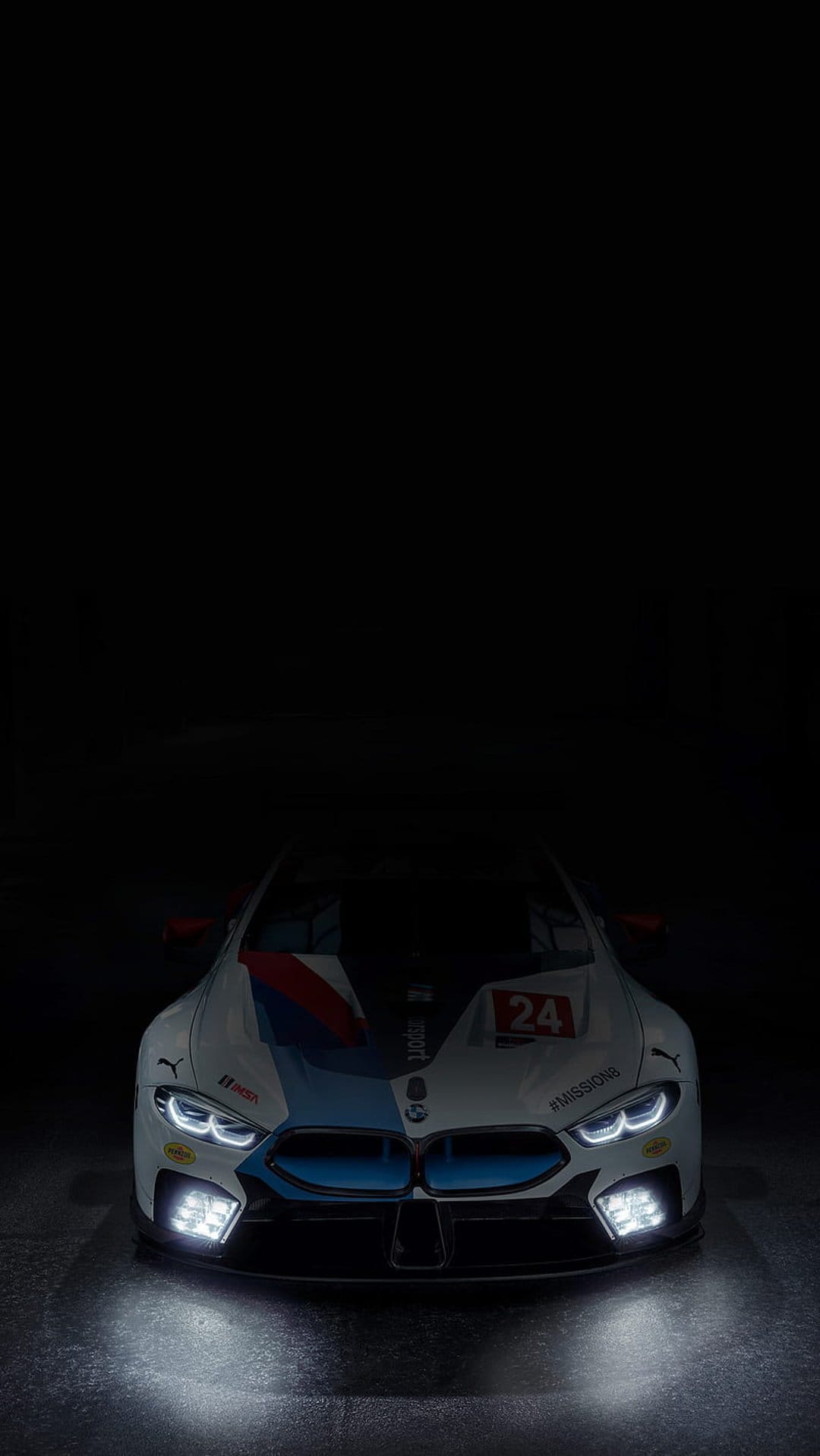 BMW M8 GTE race car with illuminated LED headlights and number 24 decals on a polished dark studio floor.