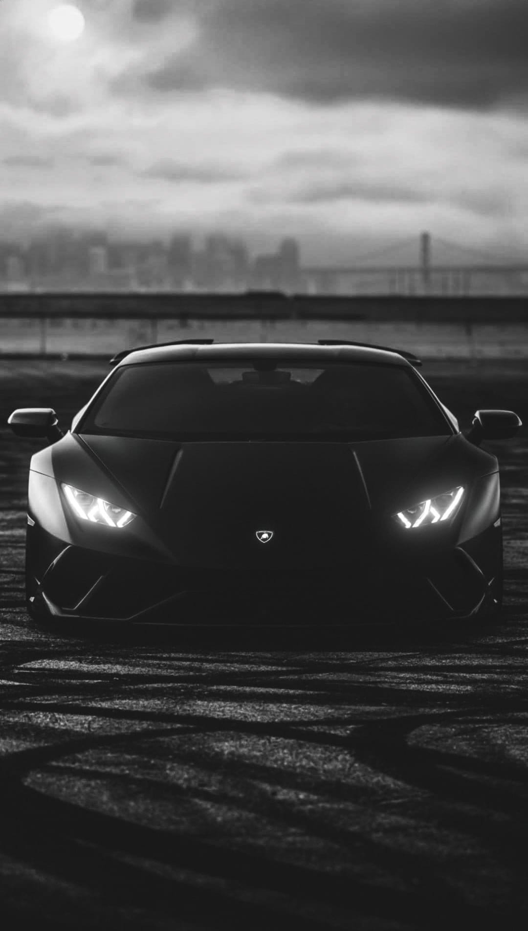 Black Lamborghini with glowing headlights on a textured road with a bridge and misty city skyline behind.