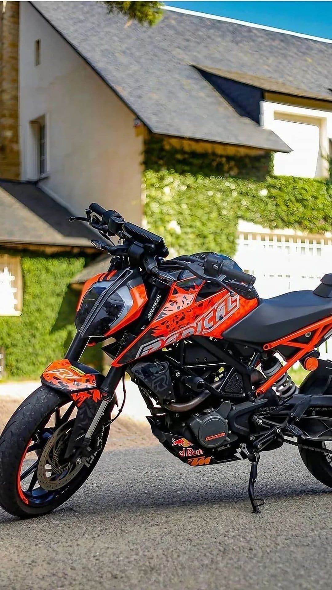 Orange KTM Duke motorcycle with radical graphics parked on asphalt before a house covered in green climbing ivy.