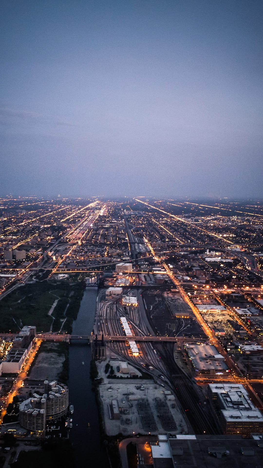Glowing golden street grids illuminate a dark urban landscape from above near a river and railway yard at night.