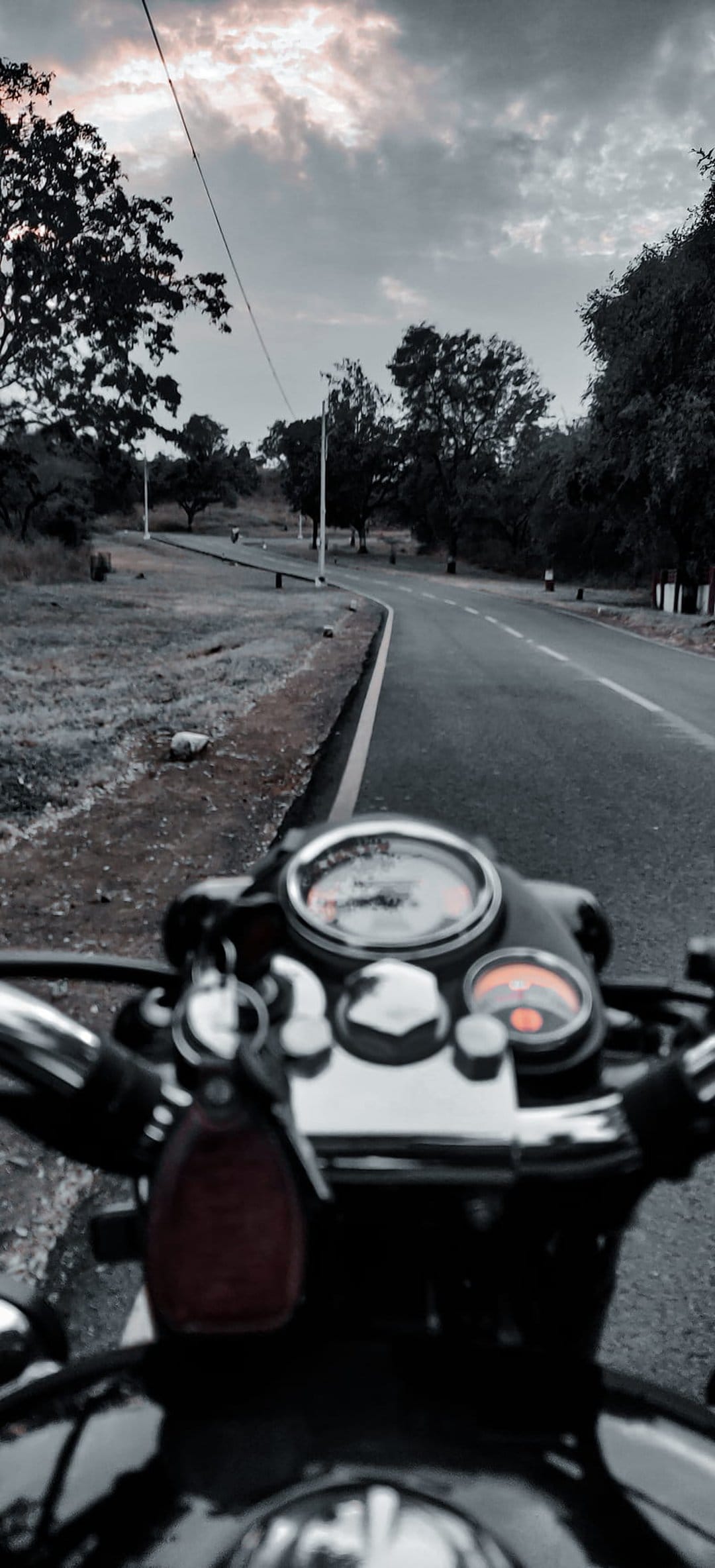 Chrome motorcycle handlebars and analog gauges overlook a curved asphalt road lined with trees beneath storm clouds.