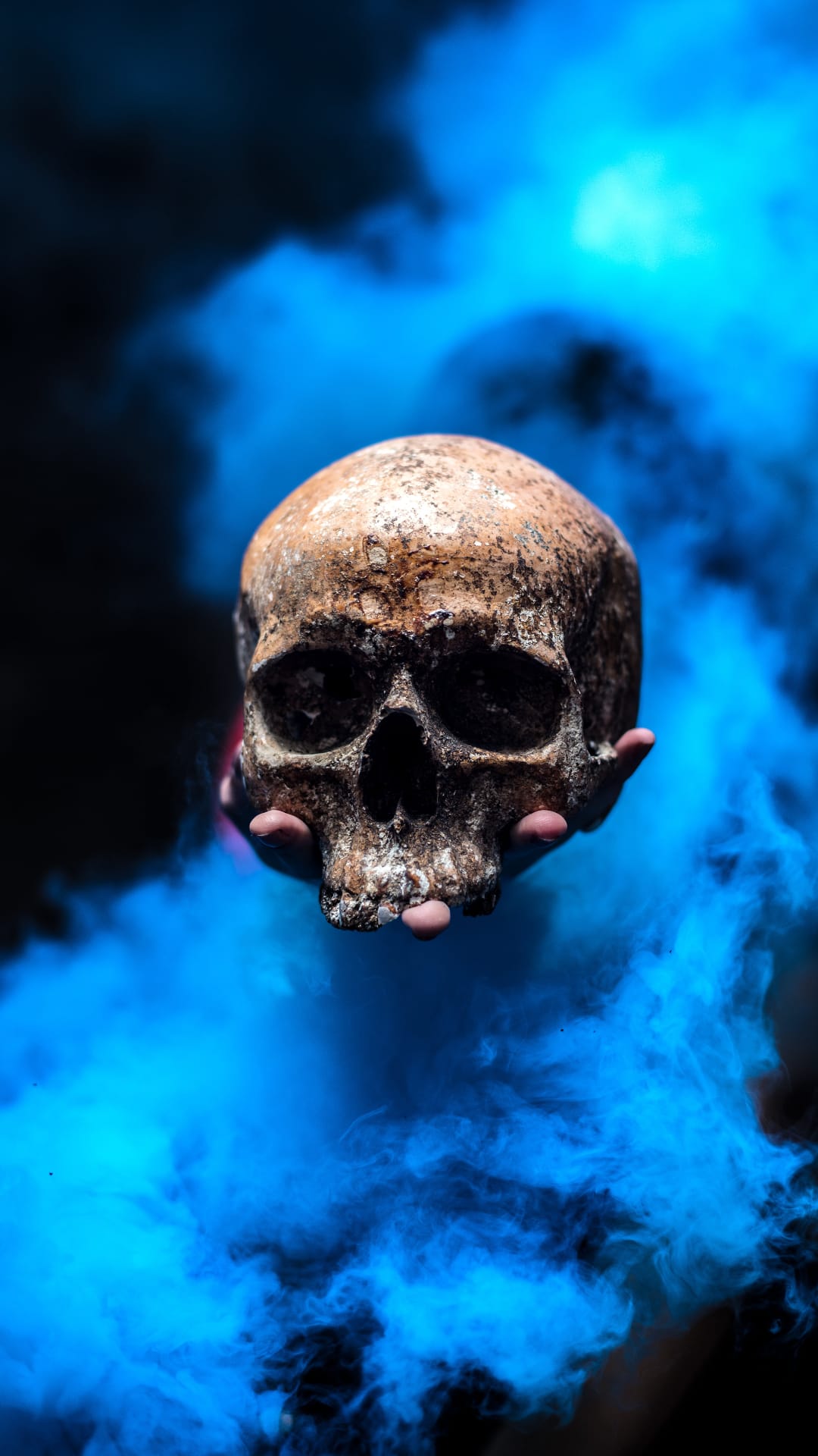 Weathered human skull held by hands within swirling neon blue smoke and ethereal vapor against a black background.