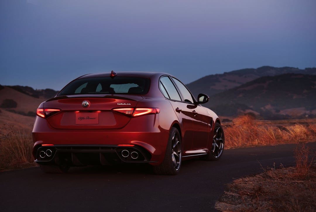Rear view of a red Alfa Romeo Giulia on a road at dusk with glowing taillights and mountain silhouettes.