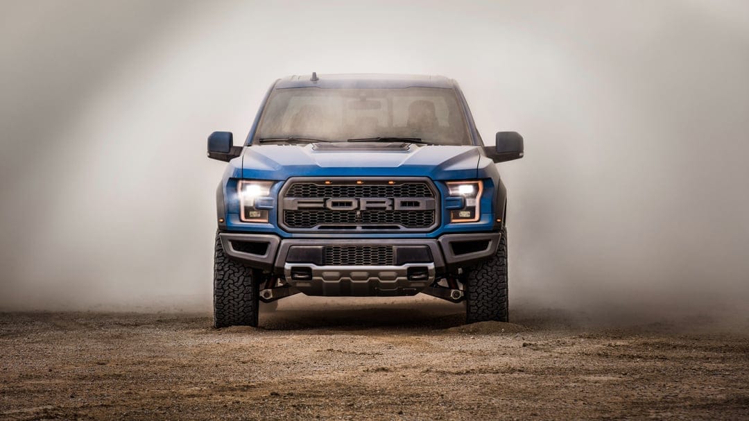 Blue Ford Raptor truck centered in a dense cloud of dust on a dirt road with headlights illuminated.