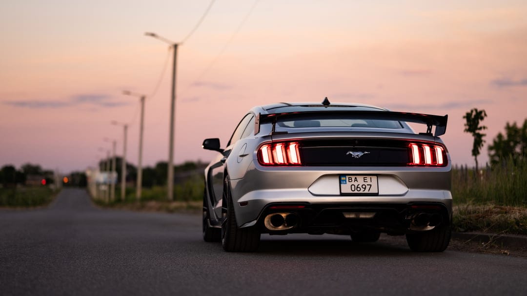 Silver Ford Mustang GT rear view with glowing taillights parked on a quiet road during a vibrant pink sunset.
